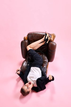 Photo for Businesswoman lying on fallen armchair, using mobile phone and shouting in anger nad irritation against pink studio background. Concept of business, working routine, deadlines, freelance, office, ad - Royalty Free Image