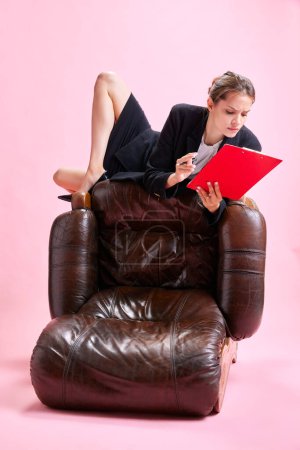 Foto de Mujer de negocios acostada en la parte superior del sillón y mirando a través de documentos sobre fondo de estudio rosa. Concepto de negocio, rutina de trabajo, plazos, freelance, oficina, anuncio - Imagen libre de derechos