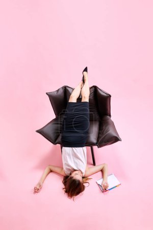 Téléchargez les photos : Femme d'affaires inconfortablement couchée sur un fauteuil, travaillant, prenant des notes sur fond de studio rose. Concept d'entreprise, routine de travail, délais, freelance, bureau, annonce - en image libre de droit
