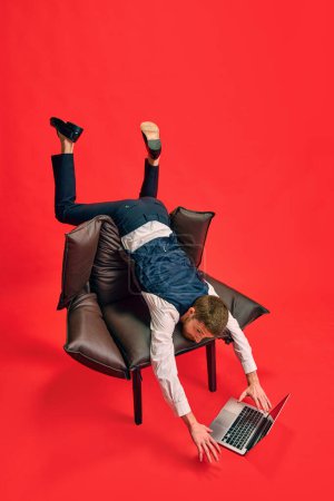 Photo for Finishing tasks. Businessman lying on armchair in weird pose, working on laptop against red studio background. Concept of business, working routine, deadlines, freelance, office, ad - Royalty Free Image