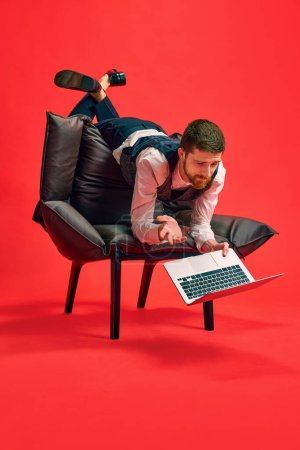 Téléchargez les photos : Finition des tâches. Homme d'affaires allongé sur un fauteuil dans une pose bizarre, travaillant sur un ordinateur portable sur fond de studio rouge. Concept d'entreprise, routine de travail, délais, freelance, bureau, annonce - en image libre de droit