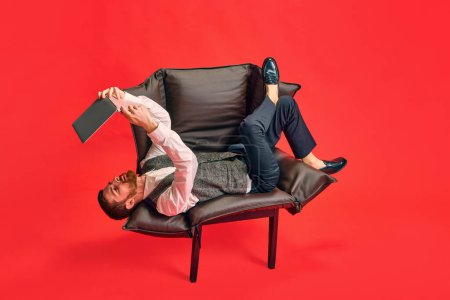 Téléchargez les photos : Homme d'affaires élégant allongé sur un fauteuil, tenant un ordinateur portable au-dessus et travaillant dans une position bizarre sur fond de studio rouge. Concept d'entreprise, routine de travail, délais, freelance, bureau, annonce - en image libre de droit