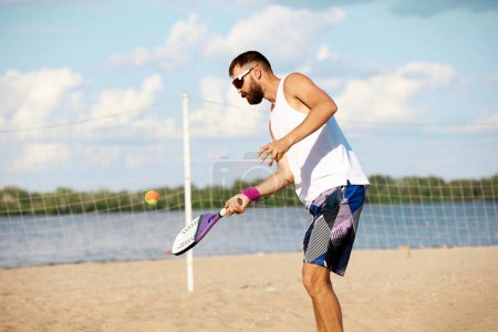 Téléchargez les photos : Jeune homme musclé en lunettes de soleil debout sur la plage près du filet et jouant au tennis en plein air par une chaude journée ensoleillée. Concept de sport, temps libre, mode de vie actif, passe-temps, jeu, été, annonce - en image libre de droit