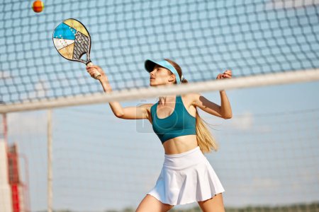 Téléchargez les photos : Jeune femme sportive jouant à la plage, au paddle tennis, frappant balle avec raquette. Entraînement en plein air par une chaude journée d'été. Concept de sport, temps libre, mode de vie actif, passe-temps, jeu, été, annonce - en image libre de droit
