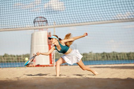 Téléchargez les photos : Femme compétitive, athlète, joueuse de tennis professionnelle en mouvement pendant le match, frappant la balle avec la raquette, s'entraînant sur la plage le jour d'été. Concept de sport, temps libre, mode de vie actif, passe-temps, publicité - en image libre de droit