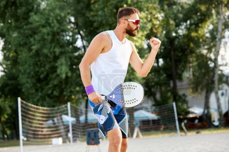 Téléchargez les photos : Homme barbu en lunettes de soleil allant sur la plage pour jouer au paddle tennis par une chaude journée d'été. Activité de plein air. Concept de sport, temps libre, mode de vie actif, passe-temps, jeu, été, annonce - en image libre de droit