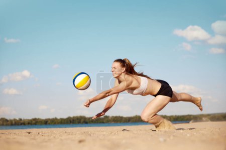 Téléchargez les photos : Jeune athlète compétitive jouant au beach-volley, frappant la balle avec les mains et tombant sur le sable. Correspondance. Concept de sport, mode de vie actif et sain, passe-temps, été, publicité - en image libre de droit