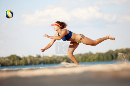 Téléchargez les photos : Image dynamique de la jeune femme en mouvement, jouant au beach-volley, frappant la balle et tombant sur le sable. Concept de sport, mode de vie actif et sain, passe-temps, été, publicité - en image libre de droit