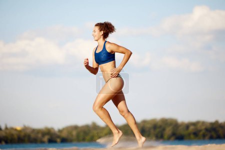 Téléchargez les photos : Jeune femme mince, en forme, corps sain courant sur la plage par une chaude journée d'été à l'extérieur. Concept de sport, mode de vie actif et sain, passe-temps, été, publicité - en image libre de droit