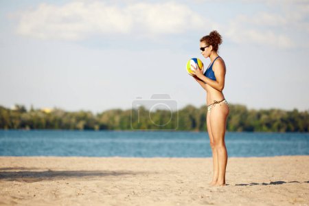Téléchargez les photos : Jeune femme sportive et sportive en maillot de bain jouant au beach-volley en plein air sur du sable près de la rivière. Air frais et entraînement. Concept de sport, mode de vie actif et sain, passe-temps, été, publicité - en image libre de droit