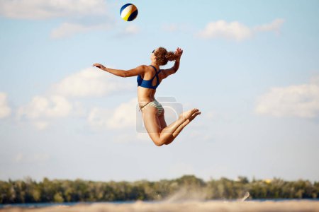 Téléchargez les photos : Jeune femme sportive et sportive en maillot de bain jouant au beach-volley en plein air sur du sable près de la rivière. Air frais et entraînement. Concept de sport, mode de vie actif et sain, passe-temps, été, publicité - en image libre de droit