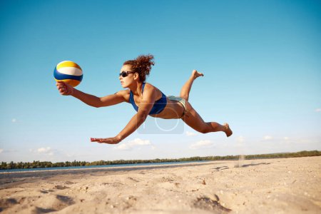 Téléchargez les photos : Image dynamique de la jeune femme en mouvement, jouant au beach-volley, frappant la balle et tombant sur le sable. Journée chaude ensoleillée. Concept de sport, mode de vie actif et sain, passe-temps, été, publicité - en image libre de droit