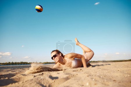 Téléchargez les photos : Image dynamique de la jeune femme en mouvement, jouant au beach-volley, frappant la balle et tombant sur le sable. Concept de sport, mode de vie actif et sain, passe-temps, été, publicité - en image libre de droit