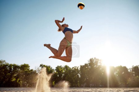 Téléchargez les photos : Image dynamique de la jeune femme sportive jouant au beach-volley, frappant la balle dans un saut par-dessus le ciel. Concept de sport, mode de vie actif et sain, passe-temps, été, publicité - en image libre de droit