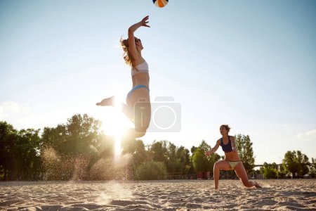 Téléchargez les photos : Deux femmes, amies jouant au beach-volley en plein air par une chaude journée d'été au coucher du soleil. Passer du temps de manière active. Concept de sport, mode de vie actif et sain, passe-temps, été, publicité - en image libre de droit