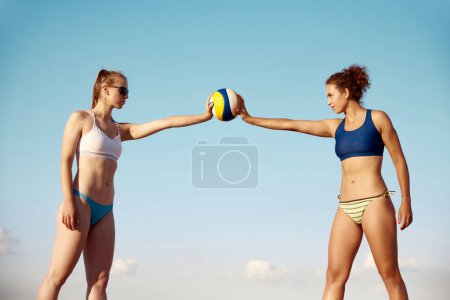 Téléchargez les photos : Deux jeunes femmes sportives, avec ajustement, corps mince en maillot de bain tenant ballon de volley-ball sur fond de ciel bleu. Concept de sport, mode de vie actif et sain, passe-temps, été, publicité - en image libre de droit
