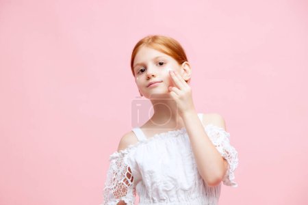 Photo for Portrait of little redhead girl, child applying face moisturizing cream against pink studio background. Concept of skincare, childhood, cosmetology and health, beauty, organic products, ad - Royalty Free Image