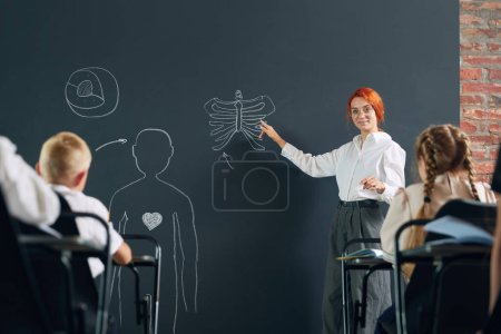 Photo for Young redhead woman, teacher standing at chalkboard and teaching anatomy to little children in primary school. Human structure. Concept of school, education, childhood, knowledge, lifestyle - Royalty Free Image