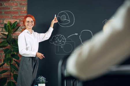 Photo for Young redhead woman, teacher standing at chalkboard and teaching anatomy to little children in primary school. Drawn elements. Concept of school, education, childhood, knowledge, lifestyle - Royalty Free Image