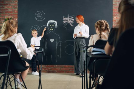 Photo for Boy, student of elementary school standing at chalkboard with teacher, answering, drawing human structure and showing to classmates. Concept of school, education, childhood, knowledge, lifestyle - Royalty Free Image