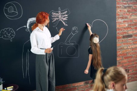Photo for Little girl, child drawing on blackboard giant heart shaped, standing with teacher and learning anatomy of human. Primary school. Concept of school, education, childhood, knowledge, lifestyle - Royalty Free Image