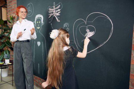 Téléchargez les photos : Petite fille, enfant dessinant sur tableau noir géant en forme de coeur, debout avec professeur et l'anatomie d'apprentissage de l'humain. L'école primaire. Concept d'école, éducation, enfance, connaissance, mode de vie - en image libre de droit