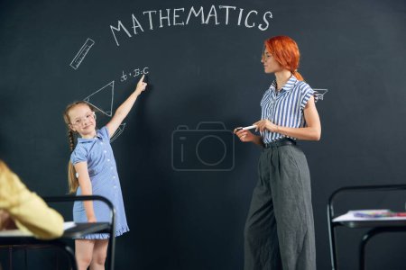 Foto de Niña, niña parada en la pizarra, dibujando triángulo, haciendo tareas matemáticas con el maestro. Geometría de aprendizaje en la escuela. Concepto de escuela, educación, infancia, conocimiento, estilo de vida - Imagen libre de derechos