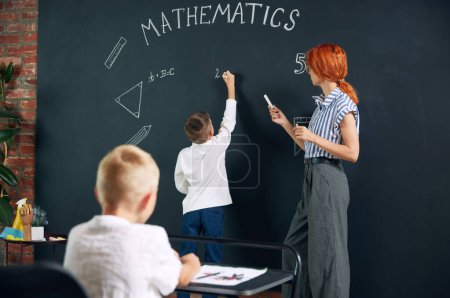 Photo for Little bot, child, student standing at chalkboard, writing and solving math tasks with teacher. Lessons at school. Concept of school, education, childhood, knowledge, lifestyle - Royalty Free Image
