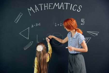 Photo for Little smart girl standing by chalkboard with teacher, writing and solving math tasks. Geometry lessons at school. Concept of school, education, childhood, knowledge, lifestyle - Royalty Free Image