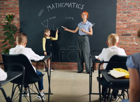 Photo for Young woman, teacher standing by blackboard with little girl, child and teaching math at school. Attentive kids listening. Concept of school, education, childhood, knowledge, lifestyle - Royalty Free Image