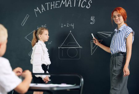 Photo for Young woman, teacher of math standing by blackboard with little girl student and solving geometry tasks. Lesson. Concept of school, education, childhood, knowledge, lifestyle - Royalty Free Image
