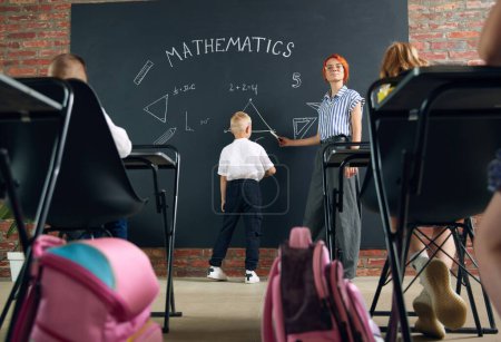 Photo for Classroom with kids learning geometry. Little boy standing by blackboard with teacher and solving math tasks. Concept of school, education, childhood, knowledge, lifestyle - Royalty Free Image