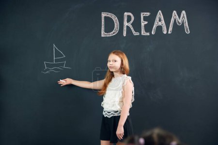 Photo for Little girl, child standing at chalkboard and drawing her dreamings. Imagination of children. Psychology and creative thinking. Concept of school, education, childhood, knowledge, lifestyle - Royalty Free Image