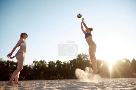Téléchargez les photos : Deux femmes, amies jouant au beach-volley en plein air par une chaude journée d'été au coucher du soleil. Passer du temps de manière active. Concept de sport, mode de vie actif et sain, passe-temps, été, publicité - en image libre de droit
