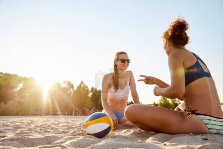 Téléchargez les photos : Deux belle jeune femme assise sur la plage au coucher du soleil, parlant joyeusement, se reposant après le match de volley-ball. Concept de sport, mode de vie actif et sain, passe-temps, été, publicité - en image libre de droit
