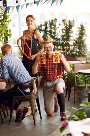 Photo for Young stylish people in bavarian clothes drinking beer and eating lager beer at pub. Festival, celebration and party. Concept of oktoberfest, traditional taste, friendship, leisure time, enjoyment - Royalty Free Image