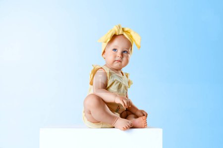 Photo for Beautiful baby girl, toddler with blue eyes, wearing yellow clothes and accessories, sitting against blue studio background. Concept of childhood, family, lifestyle, happiness, care. Copy space for ad - Royalty Free Image