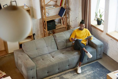Téléchargez les photos : Image vue du dessus du jeune homme africain en pull jaune, assis sur un canapé dans le salon à la maison et étudiant, travaillant sur une tablette. Concept d'entreprise et d'éducation, travail indépendant, mode de vie moderne - en image libre de droit