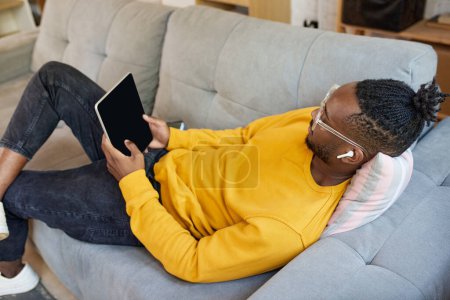 Téléchargez les photos : Jeune homme africain allongé sur le canapé, écoutant des leçons vidéo sur tablette via un casque. Travail en ligne et leçons. Concept d'entreprise et d'éducation, travail indépendant, mode de vie moderne - en image libre de droit