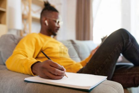 Photo for Focus on male hand making notes on notebook. Young african man sitting on sofa at home in earphones, looking on tablet and studying. Concept of business and education, freelance job, modern lifestyle - Royalty Free Image