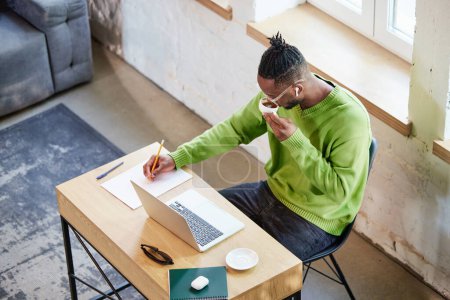 Photo for Top view image of young african man sitting at home at table, drinking coffee, working online, making notes. Concept of business and education, freelance job, modern lifestyle - Royalty Free Image