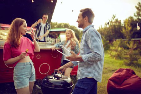 Photo for Young smiling man and woman cooking meat, doing barbecue, drinking cocktails and talking. Blurred people on background. Good time outdoors. Concept of friendship, leisure time, weekends, summer, party - Royalty Free Image