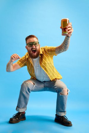 Photo for Portrait of emotional and excited bearded man in sunglasses and yellow shirt posing with beer against blue studio background. Concept of emotions, leisure time, positivity, party and celebration, ad - Royalty Free Image