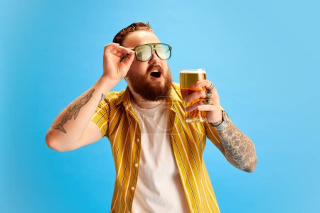 Téléchargez les photos : Homme gai barbu en chemise jaune et lunettes de soleil buvant de la bière sur fond bleu studio. Vacances et détente. Concept d'émotions, temps libre, positivité, fête et célébration, publicité - en image libre de droit