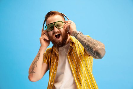 Photo for Cheerful and positive bearded man in sunglasses and yellow shirt emotionally listening to music in headphones against blue studio background. Concept of emotions, leisure time, positivity, ad - Royalty Free Image