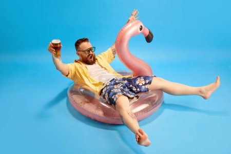Photo for Happy and relaxed bearded man in swim shorts and yellow shirt sitting in swimming circle with beer against blue studio background. Concept of emotions, leisure time, positivity, travelling, ad - Royalty Free Image