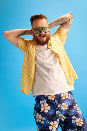 Téléchargez les photos : Homme barbu heureux, souriant et détendu en lunettes de soleil, short de bain et chemise jaune posant sur fond de studio bleu. Vacances d'été. Concept d'émotions, temps libre, positivité, publicité - en image libre de droit
