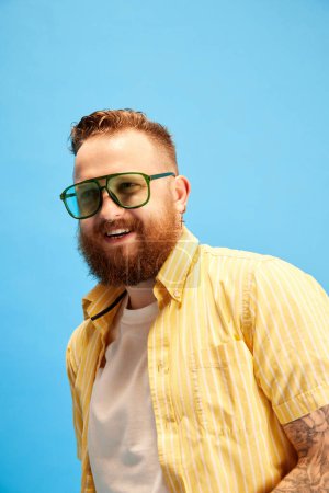 Photo for Portrait of bearded redhead man in sunglasses, wearing yellow shirt, posing against blue studio background. Concept of emotions, leisure time, vacation, summer, positivity, ad - Royalty Free Image