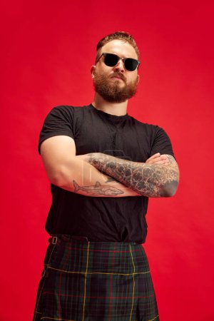 Foto de Retrato de hombre serio barbudo en gafas de sol posando en falda escocesa - escocés sobre fondo rojo estudio. Una mirada extraordinaria. Concepto de estilo de vida, estilo escocés, moda, música, diversión y alegría, anuncio - Imagen libre de derechos