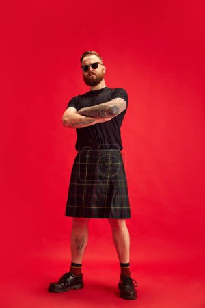 Téléchargez les photos : Portrait d'homme sérieux barbu en lunettes de soleil posant en jupe écossaise - kilt sur fond de studio rouge. Un look extraordinaire. Concept de style de vie, style écossais, mode, musique, plaisir et joie, annonce - en image libre de droit